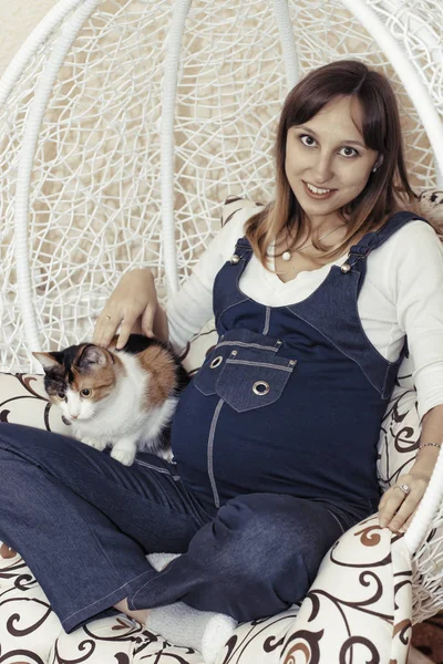 Jeune femme enceinte en jeans salopettes posant dans une chaise suspendue avec son chat — Photo