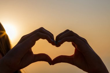 Gün batımının bulanık arka planında kalp yapan kadın eller