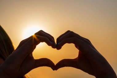 Gün batımının bulanık arka planında kalp yapan kadın eller