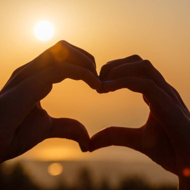 Gün batımının bulanık arka planında kalp yapan kadın eller