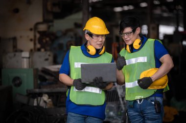Sanayi işçisi teftiş altında ve fabrika istasyonunda dizüstü bilgisayarla üretim sürecini kontrol ediyor. Mühendis iş yerinde günlük üniforma ve güvenlik kaskı giyiyor.
