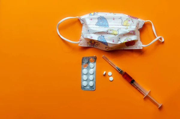 Medizinische Kindergesichtsmaske gegen ein Virus mit einer Spritze für die Impfung, Antivirus, Coronavirus weiße Tabletten in einer Packung auf orangefarbenem Hintergrund. Stockbild