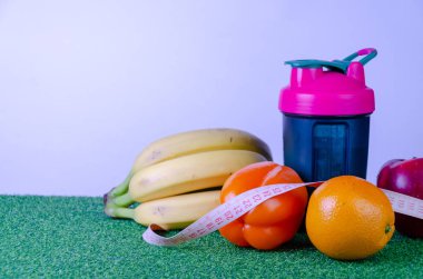 Measured blue bottle with a pink cap, a shaker for sports nutrition on green grass, measuring tape with bananas, red apple, orange bell pepper, orange on a white background, diet.