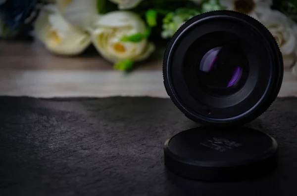 Un vecchio obiettivo per una fotocamera con una lunghezza focale di 50 mm si trova su un tavolo ed è rivolto verso la fotocamera con fiori sullo sfondo. Per fare pubblicità al fotografo . — Foto Stock