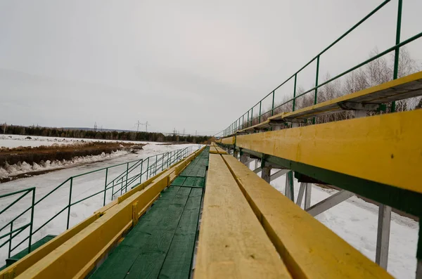 Un banco de madera amarillo y verde en un hipódromo sobre un fondo de nieve sucia en un día nublado de primavera . — Foto de Stock
