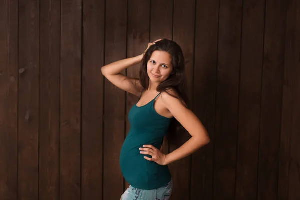 Pregnant woman in jeans shorts and green top holds hands on belly on a dark brown background. Pregnancy, maternity, preparation and expectation concept. Beautiful tender mood photo of pregnancy