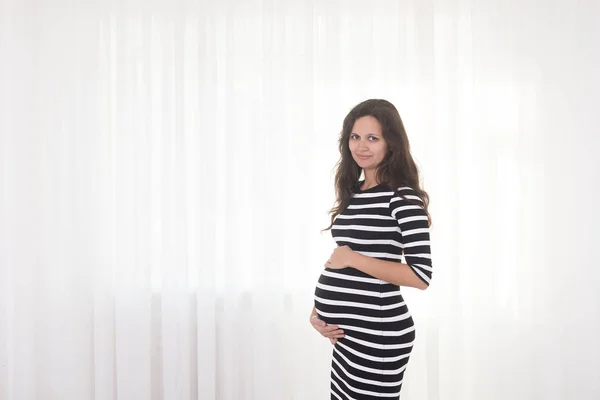 Pregnant woman in striped dress holds hands on belly on a white background. Pregnancy, maternity, preparation and expectation concept. Beautiful tender mood photo of pregnancy.