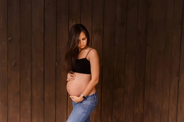 Pregnant woman in jeans and black top holds hands on belly on a dark brown background. Pregnancy, maternity, preparation and expectation concept. Beautiful tender mood photo of pregnancy