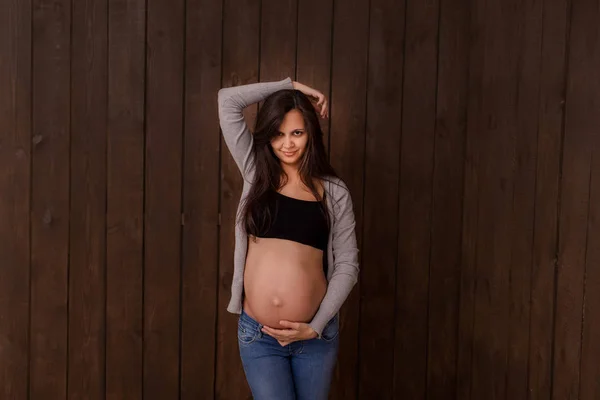 Pregnant woman in jeans and black top holds hands on belly on a dark brown background. Pregnancy, maternity, preparation and expectation concept. Beautiful tender mood photo of pregnancy — Stock Photo, Image