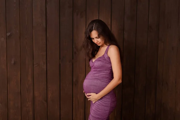Pregnant woman in violet dress holds hands on belly on a dark brown background. Pregnancy, maternity, preparation and expectation concept. Beautiful tender mood photo of pregnancy