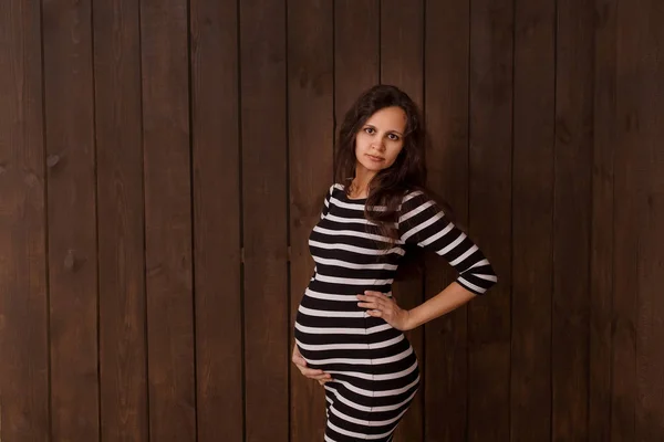 Pregnant woman in stripe dress holds hands on belly on a dark brown background. Pregnancy, maternity, preparation and expectation concept. Beautiful tender mood photo of pregnancy