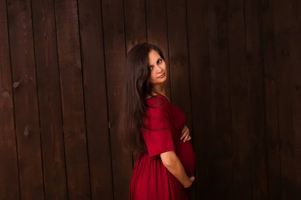 Pregnant woman in red dress holds hands on belly on a dark brown background. Pregnancy, maternity, expectation concept. Beautiful tender mood photo of pregnancy