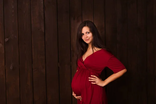 Pregnant woman in red dress holds hands on belly on a dark brown background. Pregnancy, maternity, expectation concept. Beautiful tender mood photo of pregnancy