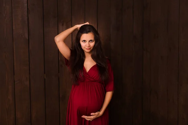 Pregnant woman in red dress holds hands on belly on a dark brown background. Pregnancy, maternity, expectation concept. Beautiful tender mood photo of pregnancy