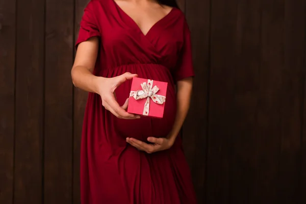Christmas pregnant woman in red dress holds in hands christmas decoration box on a dark brown background. Pregnancy, maternity, expectation concept. Beautiful tender mood photo of pregnancy