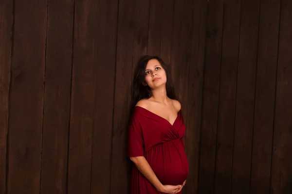 Christmas pregnant woman in red dress holds hands on belly on a dark brown background. Pregnancy, maternity, expectation concept. Beautiful tender mood photo of pregnancy