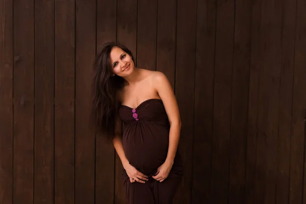 Pregnant woman in red dress holds hands on belly on a dark brown background. Pregnancy, maternity, expectation concept. Beautiful tender mood photo of pregnancy