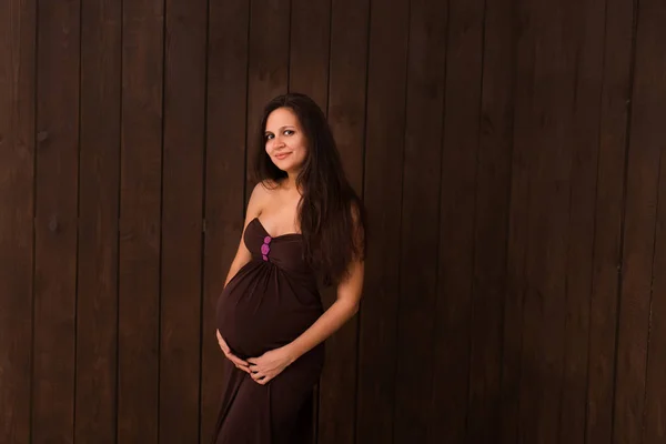 Pregnant woman in red dress holds hands on belly on a dark brown background. Pregnancy, maternity, expectation concept. Beautiful tender mood photo of pregnancy
