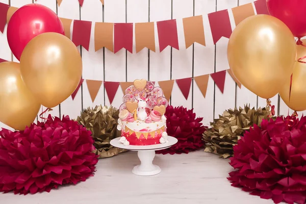 Photo zone with paper garlands, balloons, paper balls, pom poms, confetti and cream cake. Birthday cake. Smash cake. One year. Red, wine red, gold, white colors