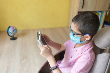 Gözlüklü çocuk, koronavirüse karşı koruyucu maske takıyor ve ödevlerini elektronik kitapla yapıyor. Karantinaya alın. Mesafe öğrenme. Uzaktan eğitim.