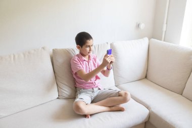 Pembe tişörtlü çocuk rahat koltukta oturur, telefon eder ve oyun oynar. Çocuk elektronik cihaz konseptini kullanıyor. Hafif arkaplan. Karantinaya alın. Tatiller.
