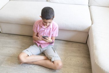 Pembe tişörtlü çocuk rahat koltukta oturur, telefon eder ve oyun oynar. Çocuk elektronik cihaz konseptini kullanıyor. Hafif arkaplan. Karantinaya alın. Tatiller.