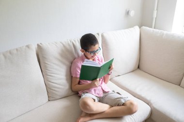 Pembe tişörtlü çocuk rahat koltukta oturup kitap okuyor. Hafif arkaplan. Karantinaya alın. Tatiller.