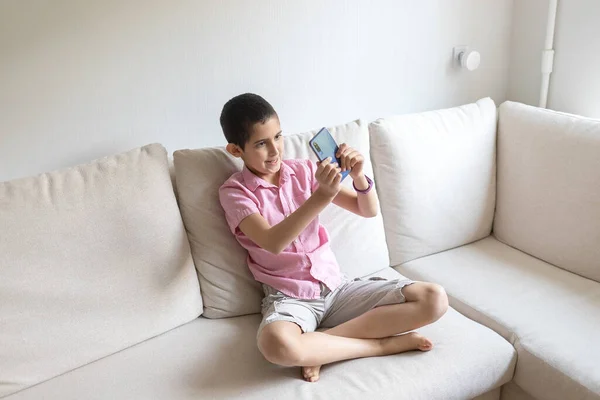 Boy in pink t-shirt sits at home on cozy sofa, uses telephone and plays. Child uses electronic devices concept. Light background. Quarantine. Holidays.