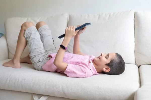 Boy in pink t-shirt lying at home on cozy sofa, uses tablet and plays. Child uses electronic devices concept. Light background. Quarantine. Holidays.