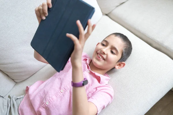 Boy in pink t-shirt lying at home on cozy sofa, uses tablet and plays. Child uses electronic devices concept. Light background. Quarantine. Holidays.
