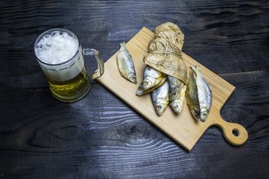 Koyu ahşap arka planda bir bardak bira, ahşap tepside kurutulmuş balık.