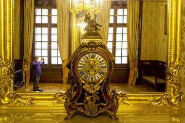 Aynada büyük altın bir saat var. Aynada küçük bir kız görünüyor. Telefondaki bir fotoğraf.