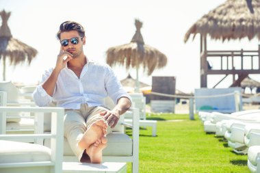 Yaz boyunca plajda beyaz sandalyelerde oturan rahat yakışıklı adam. Beyaz gömlek ve güneş gözlüğü. Bacaklarını uzat..