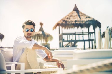 Yaz boyunca plajda beyaz sandalyelerde oturan rahat yakışıklı adam. Beyaz gömlek ve güneş gözlüğü. Arka planda ahşap kulübe.