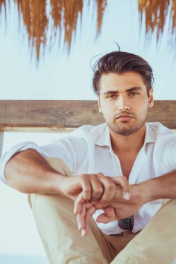 Dışarıda oturan bir adam kameraya bakıyor. Bacak bacak üstüne atmış, kollar dizlerinin üzerinde oturuyor. Moda saç ve sakal.