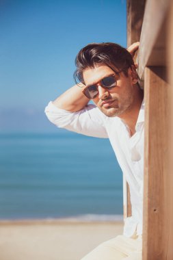 Plaj tatilinde beyaz gömlekli ve güneş gözlüklü bir adam dışarı bakıyor. Kumsalı ve mavi denizi olan deniz manzarası. Tahta bir yapının üzerinde.
