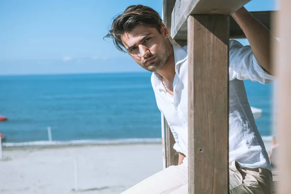 Handsome man relaxed at the sea, overlooking a wooden structure. Sea and blue sky. The young man is wearing a white shirt with beige trousers, he has black hair and a light beard.