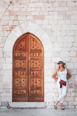 Tarihi bir kapıyla antik bir binanın cephesine yaslanmış bir kadın. Ahşap kapılı eski bir binanın tuğla duvarına yaslanan genç bir kadın. Kadın turist şapka, güneş gözlüğü ve omuz çantası takıyor..
