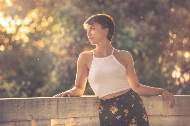 Kısa siyah saçlı, güzel, romantik genç bir kadın. Parlak, sıcak ışıklar, şehir dışında. Genç kadın beyaz bir atlet ve çiçekli bir etek giyiyor..