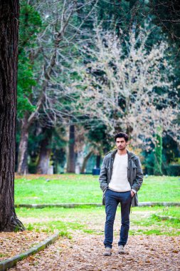 Doğal bir parkta duran yakışıklı genç adam. Kış sonbahar sahnesi, kışlık kıyafetler, ceket ve kazak. Yerde yapraklar var..