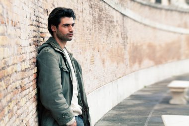 Duvara yaslanan yakışıklı bir genç adam. Günlük kıyafetler, kış tarzı. Ceket ve beyaz kazak. Sokakta, şehirde..