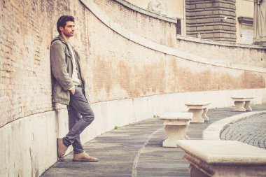 Dışarıda uzun bir duvara yaslanan yakışıklı genç adam. Günlük kıyafetler, kış tarzı. Ceket ve beyaz kazak. Sokakta, şehirde..