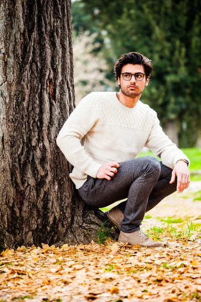 Handsome young man alone in nature near a tree outdoors. A charming young man with eyeglasses. The beautiful boy squatting on the ground leaning the tree. He dressing gowns autumn or winter.