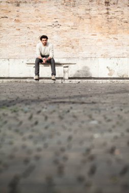 Şehirdeki caddede mermer bir bankta oturan genç adam. Ciddi tavır. Beyaz kazak. Dirsekler bacaklarda. Boşluğu kopyala.