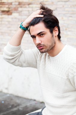 Genç yakışıklı adam elini saçlarının arasından geçiriyor. Moda saçlar ve sakal. Ciddi tavır. Beyaz kazak.