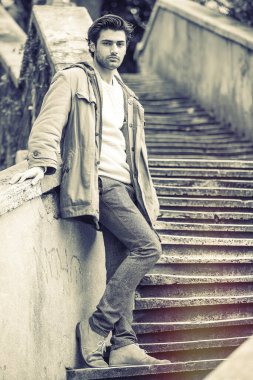 Sokakta poz veren modaya uygun genç bir adam merdivenlerde duvara yaslanıyor. Beyaz kazaklı ve ceketli kışlık giysiler. Arkasında yeşil ağaçlar.