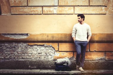 Sokakta, elleri ceplerinde, eski bir duvara yaslanmış yakışıklı genç bir adam. Moda saç stili, kendine güvenen tavırlar. Beyaz kazak. Sakal. Yanında çanta ve palto var..