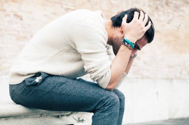 Endişeli genç adam dışarıda oturuyor ve elleri kafasında, saçları da umutsuz bir tavırla bakıyor. Beyaz kazak. Sokakta..