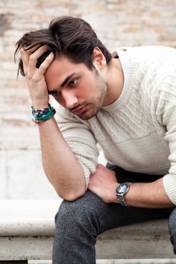 Endişeli genç adam dışarıda oturuyor ve elleri kafasında, saçları da umutsuz bir tavırla bakıyor. Beyaz kazak. Sokakta..