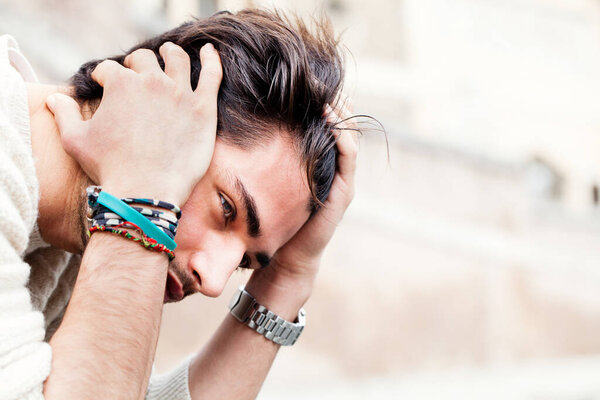 Young worried man sitting and staring outdoors with hands on the head and hair with desperate attitude. White sweater. In the street.